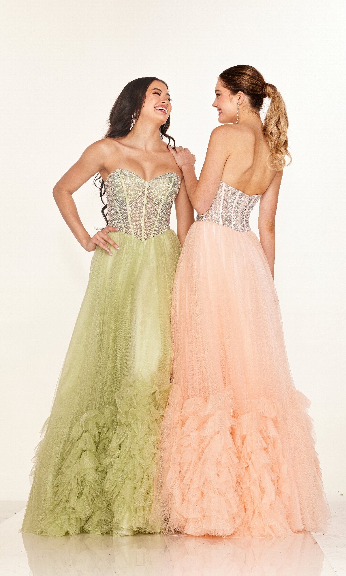 Two women wear Long Prom Dress Cecilia Couture 2617—one in pale green with an a-line tulle skirt, the other in light peach with a rhinestone corset bodice—as they stand side by side, smiling against a plain background.